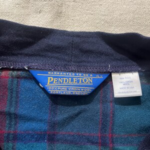 May include: Close-up of a blue, green, and red plaid shirt collar with a navy blue collar. A blue tag reads "PENDLETON 100% PURE VIRGIN WOOL PORTLAND, OREGON". A white tag reads "100% VIRGIN WOOL MADE IN USA".