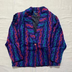 May include: A long-sleeve blazer featuring a textured, vertical wavy pattern in shades of dark blue, purple, teal, and bright pink. The garment has a wide lapel, a single front button closure, and a black interior lining with a label reading Knit Chang.