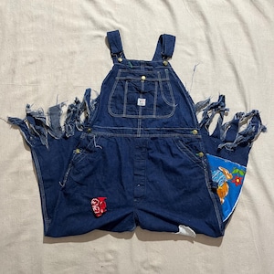 Puede incluir: Overol de mezclilla azul oscuro con bordes deshilachados y un parche rojo en la parte delantera. El overol tiene un bolsillo en el peto, bolsillos laterales y correas ajustables. Un parche azul con un conejo y flores está cosido en el bolsillo trasero.