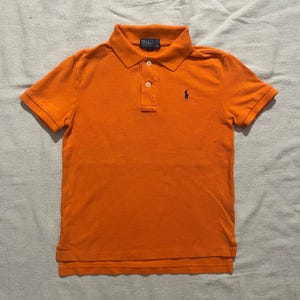 May include: Vibrant orange polo shirt with a collared neckline and short sleeves. It has a three-button placket and a navy blue logo on the chest. The shirt is made of a soft, breathable material.