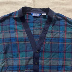 May include: A blue, green, and red plaid cardigan with a navy blue collar and trim. The cardigan has a V-neck and button closure. The brand name "Pendleton" is visible on a tag near the collar.
