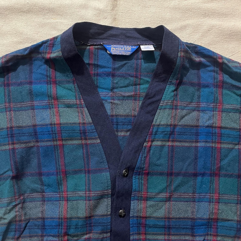 May include: A blue, green, and red plaid cardigan with a navy blue collar and trim. The cardigan has a V-neck and button closure. The brand name "Pendleton" is visible on a tag near the collar.