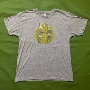 May include: A heather gray t-shirt featuring a graphic of two figures in suits playing musical instruments inside a yellow circle. One figure plays the drums, and the other plays a guitar. The background is a solid green.