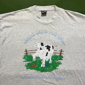 May include: A light gray t-shirt featuring a cartoon cow with black spots, standing in a grassy area next to a wooden fence. The text "Some Days You Do... Some Days You Don't" is printed above and below the cow in blue.