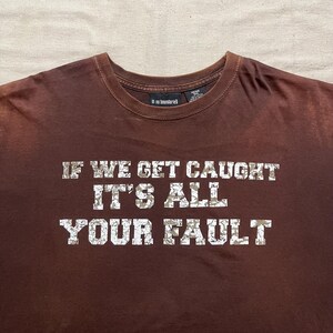 Könnte beinhalten: Braunes T-Shirt mit einem weißen, abgenutzten Aufdruck, der "Wenn wir erwischt werden, ist es deine Schuld" lautet.