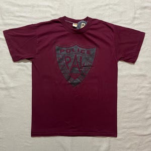 May include: Burgundy t-shirt with a black and white graphic of a shield with the text "Police Athletic League" inside.