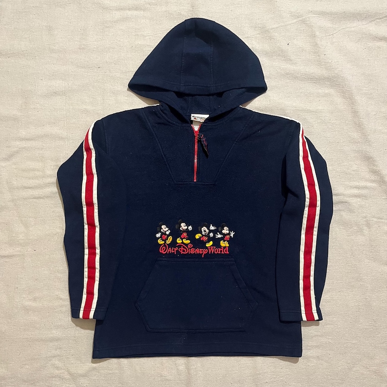 May include: A navy blue hooded sweatshirt with red and white stripes on the sleeves. The sweatshirt has a half-zip closure and a kangaroo pocket. It features an embroidered image of Mickey Mouse and the text "Walt Disney World".