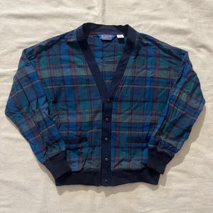 May include: A blue, green, and red plaid cardigan with a navy blue trim and button closure. The long-sleeved sweater has two front pockets and a V-neck design. The cuffs and bottom hem are navy blue.