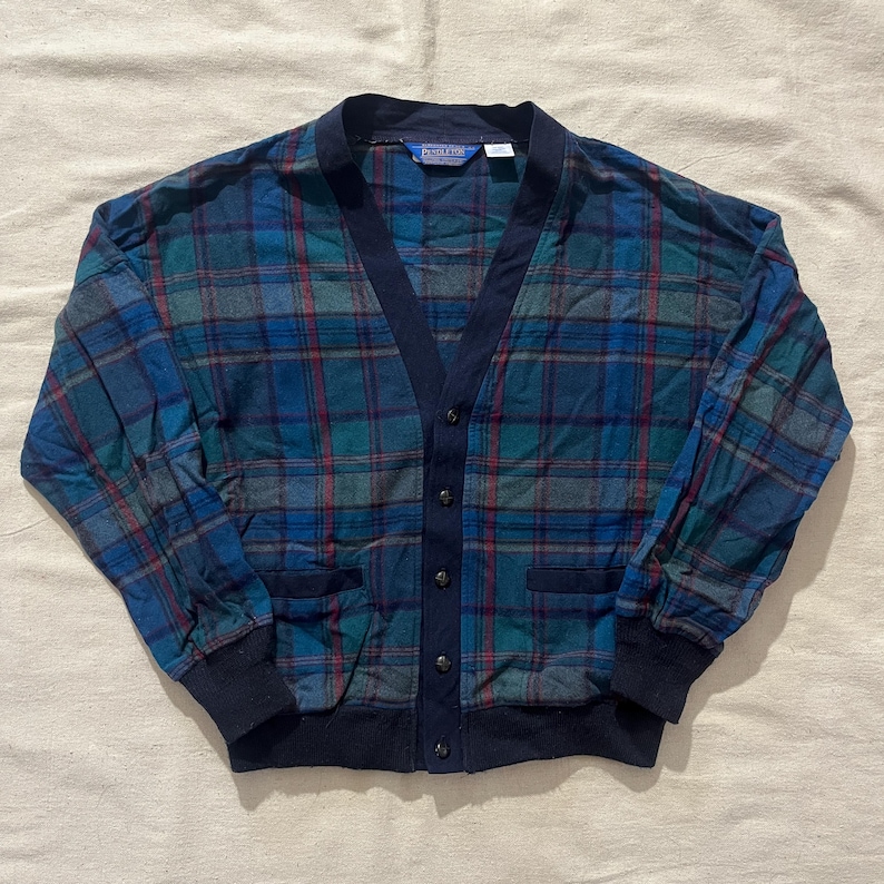 May include: A blue, green, and red plaid cardigan with a navy blue trim and button closure. The long-sleeved sweater has two front pockets and a V-neck design. The cuffs and bottom hem are navy blue.