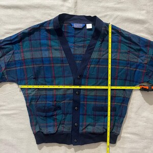 May include: A vintage Pendleton cardigan in a blue, green, and red plaid pattern. The cardigan has a navy blue trim, pockets, and buttons. The garment is being measured with a yellow tape measure. The cardigan is approximately 28 inches wide.