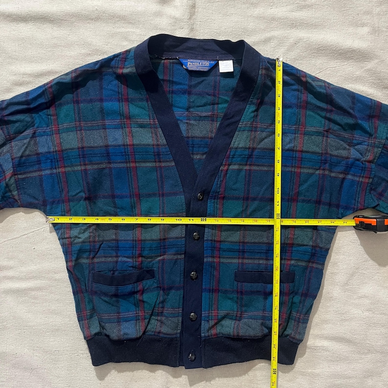 May include: A vintage Pendleton cardigan in a blue, green, and red plaid pattern. The cardigan has a navy blue trim, pockets, and buttons. The garment is being measured with a yellow tape measure. The cardigan is approximately 28 inches wide.