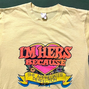 May include: A pale yellow t-shirt featuring the phrase "I'M HERS BECAUSE SHE APPRECIATES PERFECTION" in orange and pink. The design includes a pink heart and floral details. The shirt is made of a soft fabric.