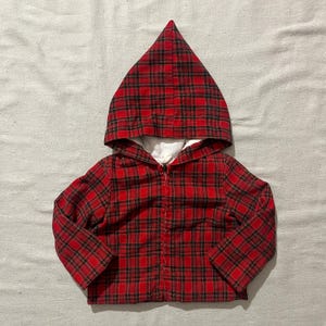 Op de afbeelding: Een rood-zwarte geruite jas met capuchon en een puntige capuchon. De jas heeft een ritssluiting over de hele lengte en is gevoerd met witte stof. Een klassieke stijl, geschikt voor diverse gelegenheden.