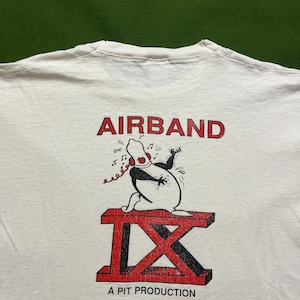 May include: White t-shirt with a red and black graphic of a cartoon character holding a phone and singing. The text "AIRBAND IX A PIT PRODUCTION" is printed below the graphic.