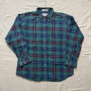 May include: A long-sleeved button-down shirt in a teal, navy, burgundy, and white plaid pattern. The shirt has a button-down collar, a chest pocket, and buttoned cuffs. The brand name is visible on the collar.