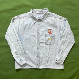 May include: Light blue denim button-down shirt with long sleeves. Features a small pocket on the chest with a cartoon character and the word "AUM" embroidered in yellow. The shirt has a classic collar and buttoned cuffs.