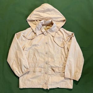 May include: A light beige hooded jacket with a button-up front, two front pockets, and a collared neckline. The jacket has long sleeves and a drawstring hood. The jacket is made of a lightweight material.