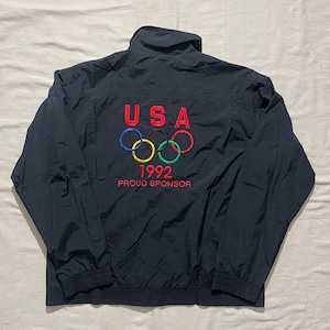May include: A navy blue windbreaker jacket featuring an embroidered design on the back. The embroidery includes the red text USA, the five interlocking Olympic rings in blue, yellow, black, green, and red, the year 1992, and the red text PROUD SPONSOR.