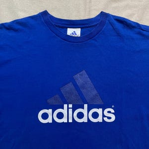 May include: Royal blue t-shirt featuring the Adidas logo in a lighter shade of blue. The Adidas logo is prominently displayed on the front of the shirt, with the brand name printed below it in white.