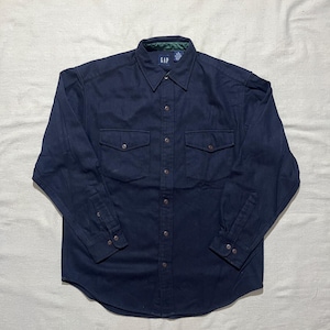 May include: A dark navy blue long-sleeved button-down shirt featuring a classic collar, two chest pockets with buttoned flaps, and a green plaid interior neck lining. The shirt includes a GAP brand label at the collar and buttoned cuffs.