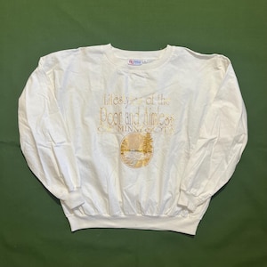 Puede incluir: Sudadera blanca de manga larga con un estampado gráfico dorado que dice "Lifestyles of the Poor and Aimless of Minnesota". El gráfico también incluye una imagen estilizada de un lago y árboles.