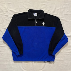 May include: A black and royal blue quarter-zip fleece pullover featuring a high collar, long sleeves with black cuffs, and a drawstring hem. A small white graphic of a hand making a shaka sign is embroidered on the left chest near the zipper.