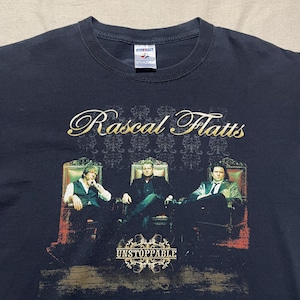 May include: A black short-sleeve t-shirt featuring a graphic print of the band Rascal Flatts. The design displays three individuals seated in ornate chairs with the text Rascal Flatts above and the word UNSTOPPABLE in gold lettering below.