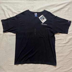 May include: A black short-sleeved t-shirt with a white Champion logo on the front. The t-shirt is laying flat on a white surface.