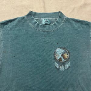 Peut inclure: T-shirt col rond de couleur teal avec un motif amérindien sur le devant. Le motif présente un emblème circulaire avec le profil d'un chef amérindien, orné d'une coiffe de plumes et de détails de capteur de rêves.