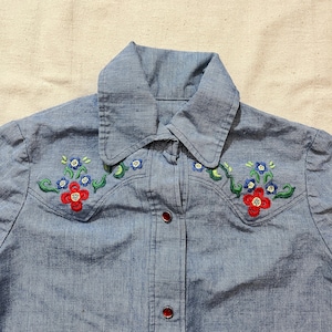 May include: A light blue denim shirt with floral embroidery. The embroidery features red, blue, and green flowers and vines. The shirt has a pointed collar and red buttons down the front.