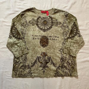May include: A long-sleeved, olive green shirt with a round neckline. The shirt features a distressed, textured pattern with ornate, dark brown floral and scroll designs. The text on the shirt reads "...kind's...develop...ent...ns...good Actions."