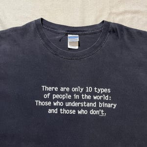 Peut inclure: Un t-shirt noir à col rond avec le texte blanc suivant : There are only 10 types of people in the world: Those who understand binary and those who don't. Le t-shirt présente une étiquette de marque Gildan Ultra Cotton à l'intérieur du col.
