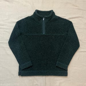 May include: A dark forest green quarter-zip pullover sweater made of a textured, fuzzy fleece material. The garment features a high collar, long sleeves with banded cuffs, and a straight hemline with a small side slit on the bottom left side.