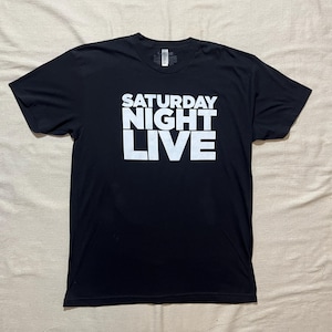 May include: A black short-sleeved t-shirt featuring the text SATURDAY NIGHT LIVE printed in large white capital letters across the center of the chest. The garment has a crew neckline and a standard fit, with a visible size label at the inner neck.