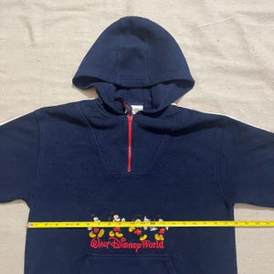 May include: A navy blue fleece pullover hoodie with a red zipper and white trim. The hoodie features a Mickey Mouse design with the text "Walt Disney World" on the front.