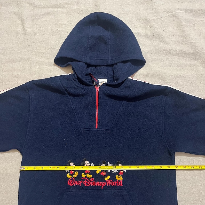 May include: A navy blue fleece pullover hoodie with a red zipper and white trim. The hoodie features a Mickey Mouse design with the text "Walt Disney World" on the front.