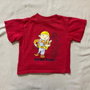 Puede incluir: Camiseta roja de manga corta con un dibujo animado de Bob el Constructor. El diseño muestra a Bob con un casco amarillo, sosteniendo planos y una caja de herramientas. El texto "Bob the Builder" está impreso debajo.