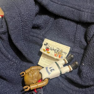 May include: A navy blue fleece hoodie with a Walt Disney World Kids tag and a Mickey Mouse logo. The hoodie has a zipper and a hood. A small toy basketball player is lying on top of the hoodie.