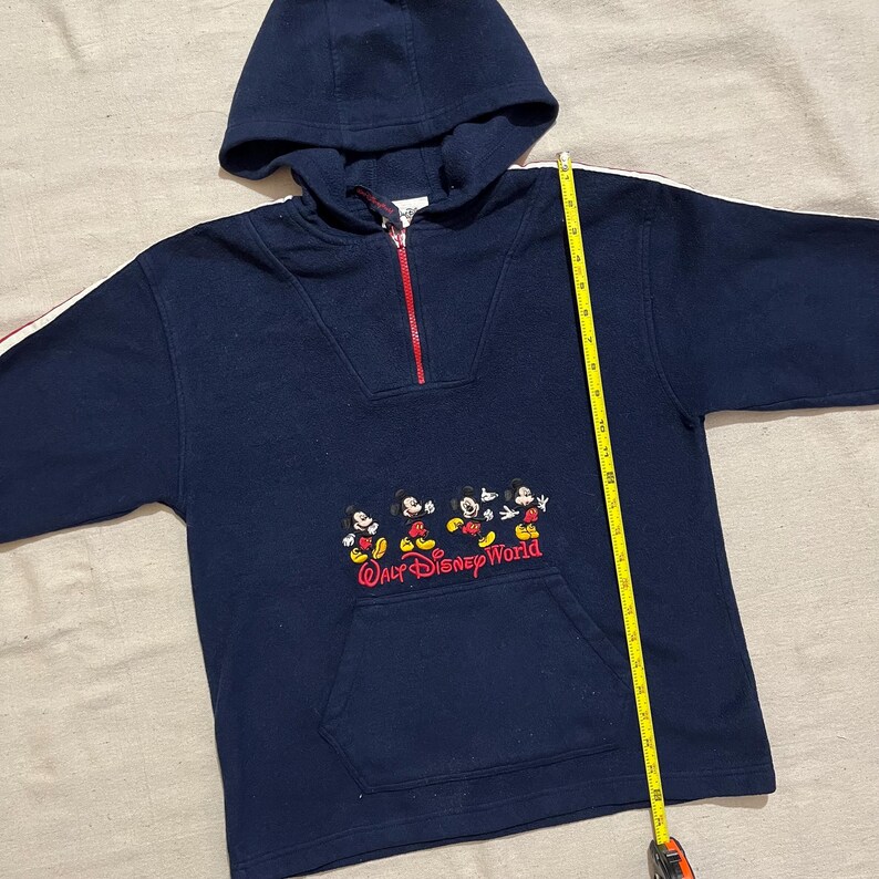 May include: A navy blue hooded sweatshirt with white and red stripes on the sleeves. It features a half-zip closure and a kangaroo pocket. The front of the sweatshirt has an embroidered image of Mickey Mouse and the text "Walt Disney World".