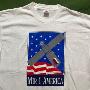 以下が含まれることがあります： 白いTシャツに、宇宙ステーションのグラフィックと「Mir 1 America」の文字、青と白のアメリカ国旗が描かれています。