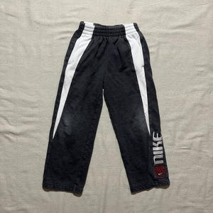 May include: Dark gray Nike sweatpants with white side stripes. The Nike logo is printed vertically on the lower leg in white, with a red basketball graphic. The pants have an elastic waistband and are likely for children.