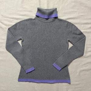 Op de afbeelding: Een grijze geribbelde coltrui met lavendelkleurige afwerking op de kraag, manchetten en zoom. De trui met lange mouwen is gemaakt van een zacht, comfortabel materiaal.