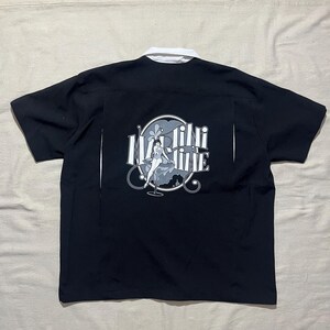 May include: A black short-sleeved button-down shirt with a white collar. The back features a graphic of a person sitting in a martini glass with the text "Martini" in a stylized font, set against a circular light blue and white background.