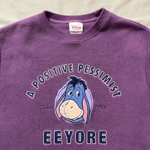 Könnte beinhalten: Lila Fleece-Sweatshirt mit Eeyore aus Winnie the Pooh. Das Design enthält den Schriftzug "A Positive Pessimist" und den Namen "Eeyore" in weißer Schrift. Das Disney-Logo ist sichtbar.