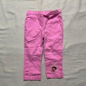 May include: Bright pink pants featuring a waistband with a single button closure, a front zipper fly, and two side pockets. The lower left leg displays an embroidered graphic of the character Dora the Explorer surrounded by flowers and three buttons.