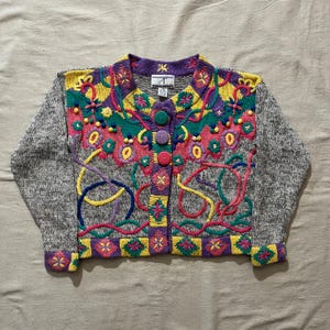 May include: A gray knit cardigan with colorful embroidered floral and abstract designs. The sweater features a button-up front with large, round buttons. The sleeves are a solid gray, while the body is adorned with pink, yellow, green, and purple details.