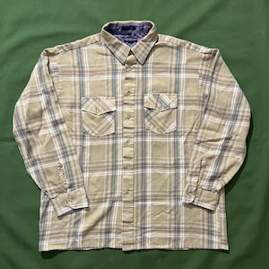 May include: A tan plaid button-down shirt with two chest pockets and long sleeves. The shirt features a classic collar and buttoned cuffs. The plaid pattern includes shades of tan, gray, and white. The shirt is made of a soft, comfortable material.