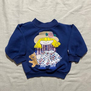 May include: A navy blue sweatshirt featuring a whimsical graphic of a girl wearing a straw hat with a pink rose and holding a teddy bear. The girl's dress is white with a floral pattern. The sweatshirt has a high neckline and long sleeves.