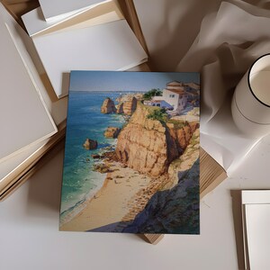May include: A watercolor print of a coastal scene featuring a beach, cliffs, and buildings. The artwork showcases a vibrant blue ocean, sandy beach, and reddish-brown cliffs. White buildings with red tile roofs are perched atop the cliffs. The print is displayed with other art supplies.
