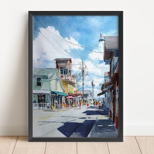 May include: Watercolor painting of a coastal street scene with buildings in shades of green, yellow, and red. A blue sky with fluffy clouds is above. People are walking on the street. The artwork is framed in black.
